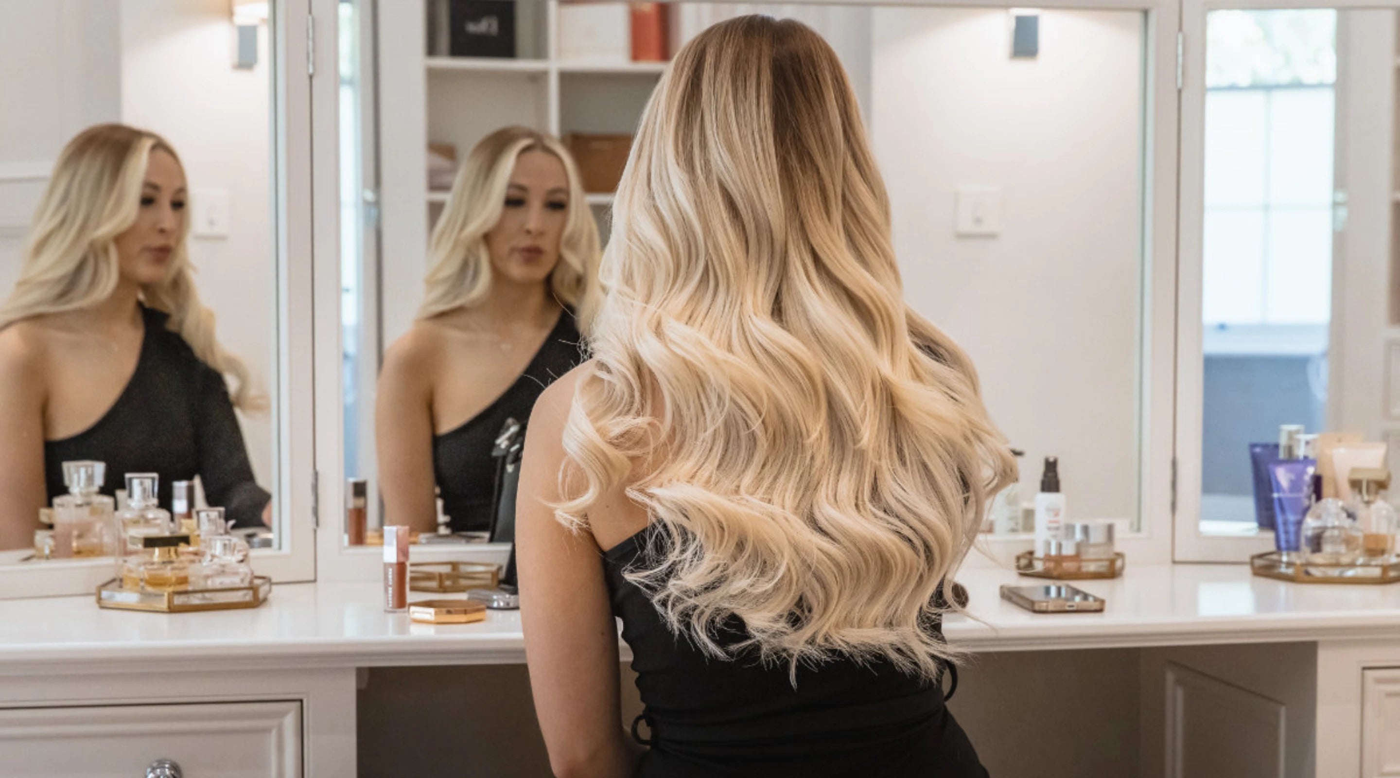 Woman with long, voluminous blonde wavy gdollshair extensions viewed from behind, standing at vanity mirror in luxury salon
