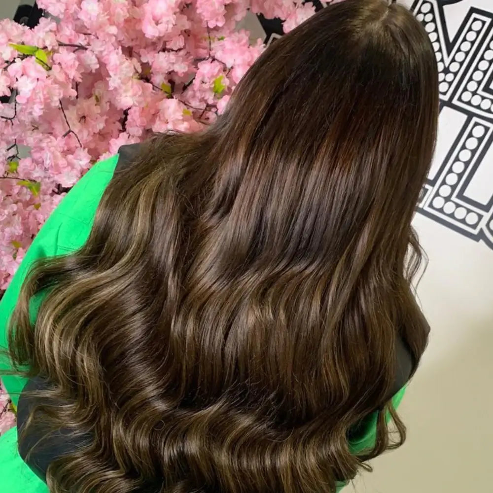 Long, wavy brown hair with golden highlights showcasing gdollshair extensions, worn with a green top against pink cherry blossoms.